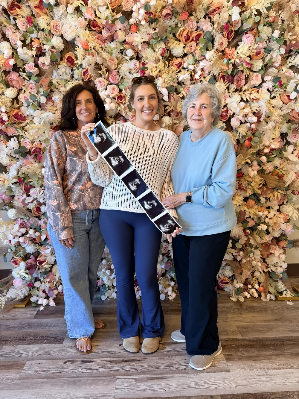 Three studio guests holding a strip of ultrasound pictures together in front of the flower wall
