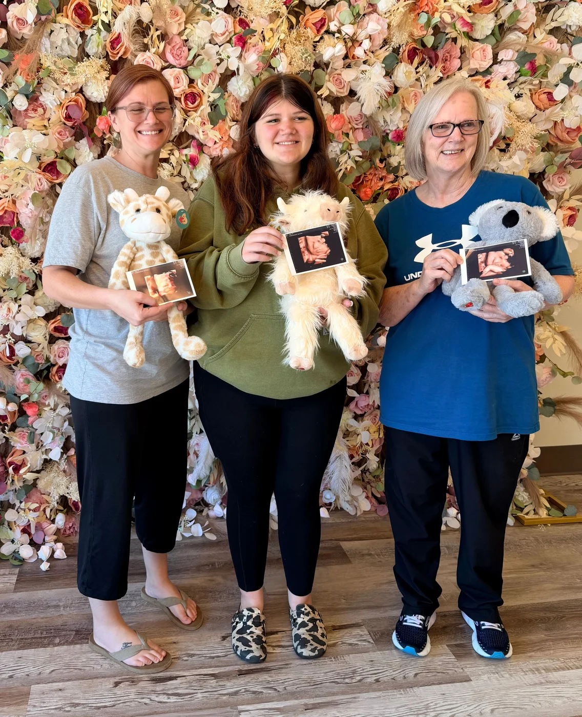 Three guests holding heartbeat keepsake animals and ultrasound prints in front of a floral wall
